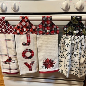 May include: Four decorative kitchen towels hanging on an oven. The towels feature various holiday patterns, including plaid, snowflakes, and the word "JOY". One towel has a cardinal, another a poinsettia, and another a green and white leaf pattern.