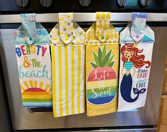 May include: Four kitchen towels hanging on a silver oven door. The towels have different designs and sayings. The first towel is blue with a beach scene and says "Beauty & the beach". The second towel is yellow with stripes and lemons. The third towel is yellow with a pineapple and says "Life is Sweet". The fourth towel is white with a mermaid and says "Lake Hair don't care".