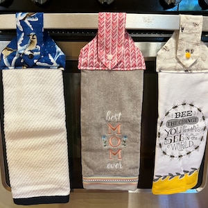 Op de afbeelding: Drie keukendoeken hangen aan een rek. De handdoek links heeft een blauwe achtergrond met een vogelpatroon. De middelste handdoek heeft een grijze achtergrond met de tekst "best mom ever" erop geborduurd. De handdoek rechts heeft een witte achtergrond met een bijenpatroon en de tekst "Bee the change you wish to see in the world".