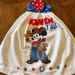 May include: White kitchen towel with a cowboy Mickey Mouse graphic. The towel features the words "Howdy Y'all" in red and blue lettering. Red and blue stars and a horseshoe are at the bottom. The top is blue and red patterned fabric.