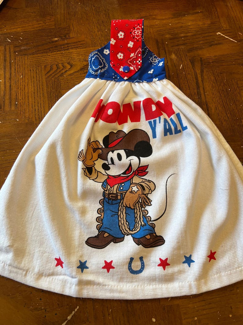 May include: White kitchen towel with a cowboy Mickey Mouse graphic. The towel features the words "Howdy Y'all" in red and blue lettering. Red and blue stars and a horseshoe are at the bottom. The top is blue and red patterned fabric.
