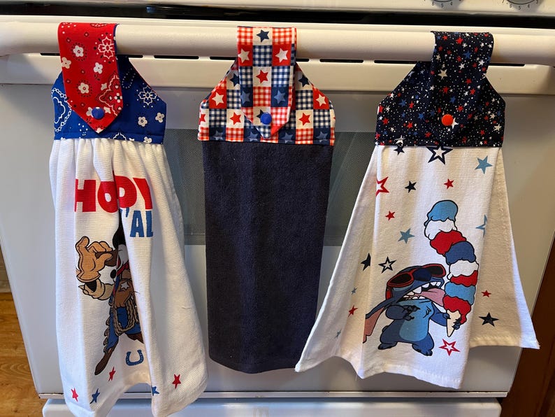 May include: Three kitchen towels with patriotic designs hanging on an oven. The towels feature red, white, and blue patterns. One towel has a cowboy graphic and the text "Howdy Y'all". Another has a Stitch graphic with an ice cream cone.