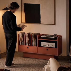 Op de afbeelding: Een terracotta-kleurig mediameubel met open verticale vakken voor het opbergen van vinylplaten en een plank voor een platenspeler en versterker. De unit bevat twee onderste lades met ronde grepen, geplaatst tussen twee houten luidsprekers.