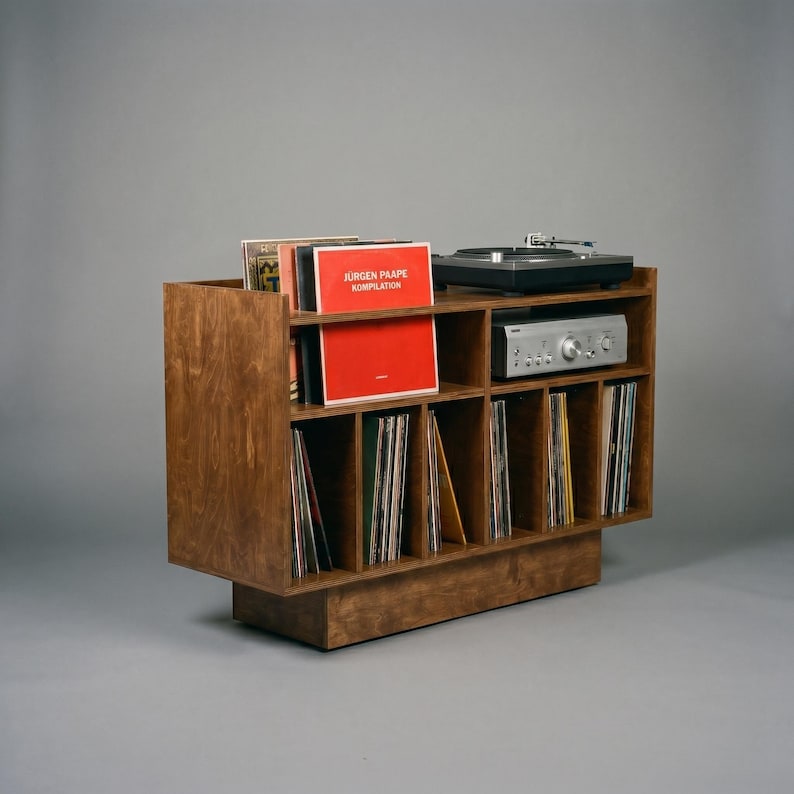 Handcrafted walnut-finish record player stand with vinyl storage, featuring a turntable on top, a stereo amplifier shelf, and four divided lower compartments filled with LP records, photographed against a neutral gray background.