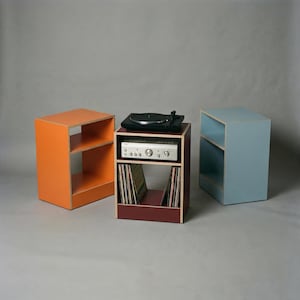 Modular plywood record player stand and vinyl storage system with natural plywood edges displayed as three separate units against a gray background. The center oxide red unit holds a black turntable on top, a silver stereo component on the middle shelf, and vinyl records below, while the orange unit on the left and chalk blue unit on the right show the modular design.