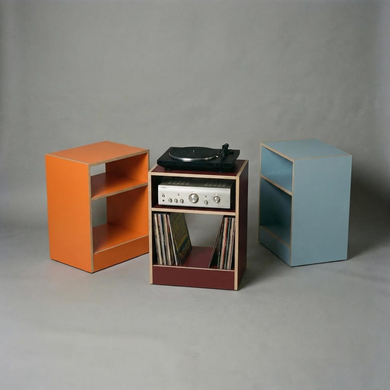 Modular plywood record player stand and vinyl storage system with natural plywood edges displayed as three separate units against a gray background. The center oxide red unit holds a black turntable on top, a silver stereo component on the middle shelf, and vinyl records below, while the orange unit on the left and chalk blue unit on the right show the modular design.