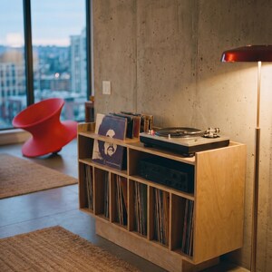 Natural birch plywood turntable console with vinyl storage, amplifier shelf, and record display beside a concrete wall in a modern apartment listening corner.