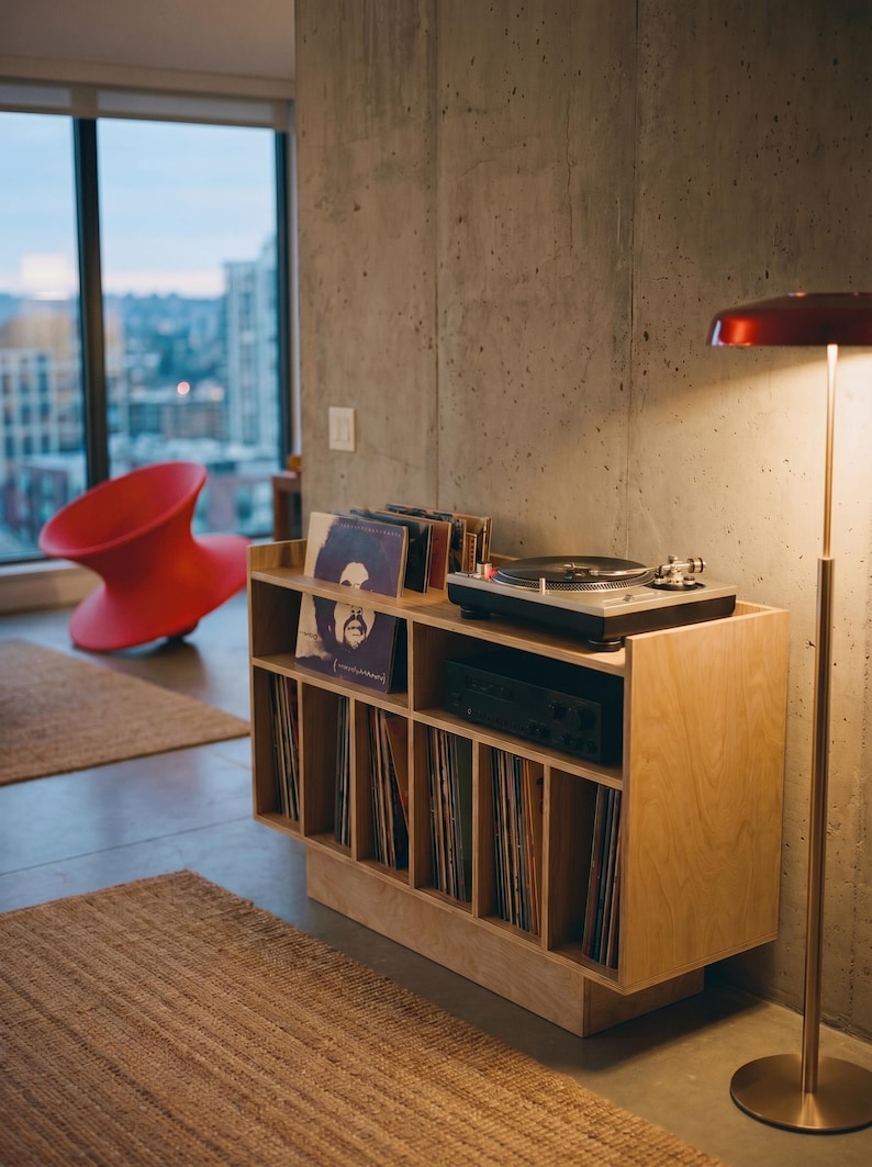 Natural birch plywood turntable console with vinyl storage, amplifier shelf, and record display beside a concrete wall in a modern apartment listening corner.