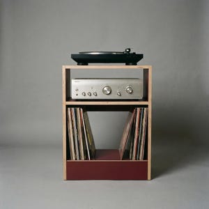 Modular plywood record player stand shown straight on against a gray background with natural plywood edges and an oxide red lower section. A black turntable sits on top with a silver stereo component on the middle shelf and vinyl records stored upright in the bottom compartment.