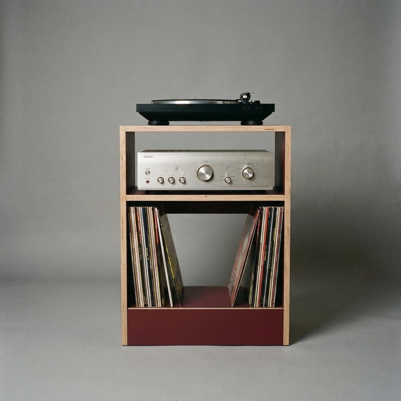 Modular plywood record player stand shown straight on against a gray background with natural plywood edges and an oxide red lower section. A black turntable sits on top with a silver stereo component on the middle shelf and vinyl records stored upright in the bottom compartment.