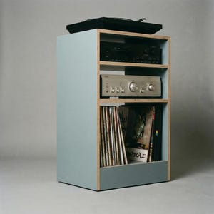 Modular plywood record player stand in chalk blue shown from an angled side view against a gray backdrop. A black turntable sits on top with two stereo components on the open shelves and vinyl records stored upright in the lower compartment, showing the slim footprint and built in storage.