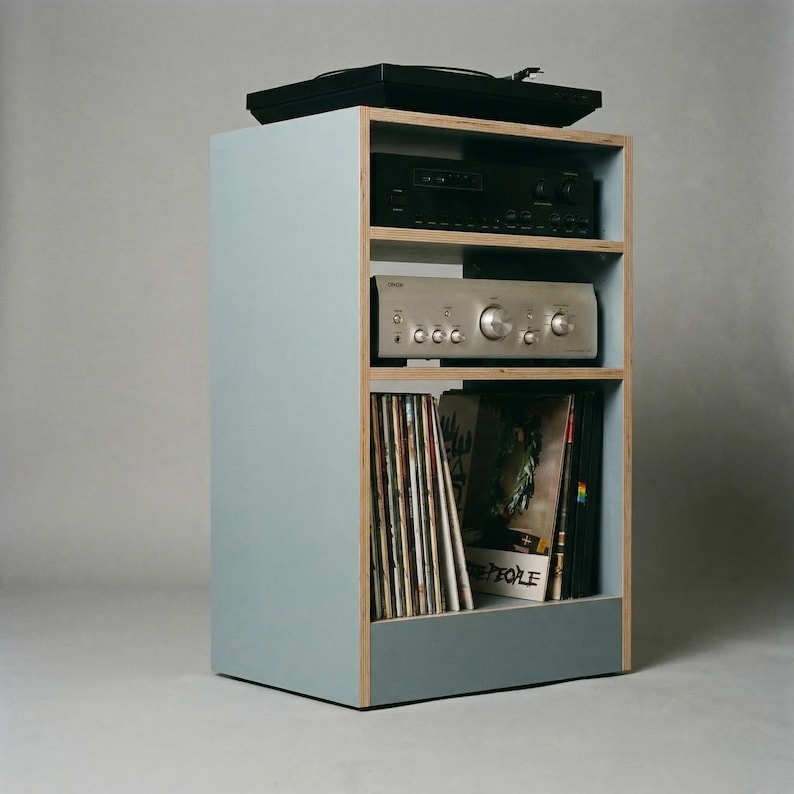 Modular plywood record player stand in chalk blue shown from an angled side view against a gray backdrop. A black turntable sits on top with two stereo components on the open shelves and vinyl records stored upright in the lower compartment, showing the slim footprint and built in storage.