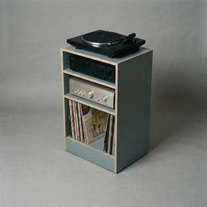 Modular plywood record player stand made from 18mm plywood in chalk blue with natural plywood edges, shown from a front angled view against a gray background. A black turntable sits on top with two stereo components on the shelves and vinyl records stored upright in the lower section.