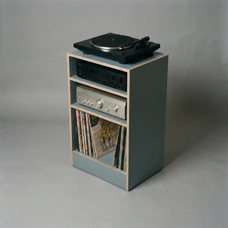 Modular plywood record player stand made from 18mm plywood in chalk blue with natural plywood edges, shown from a front angled view against a gray background. A black turntable sits on top with two stereo components on the shelves and vinyl records stored upright in the lower section.
