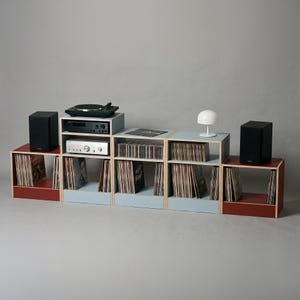 Modular plywood record player stand and vinyl storage system with natural plywood edges shown as a wide low console against a gray background. Oxide red and chalk blue modules hold a turntable and stereo components in the center with vinyl records and CDs stored across the shelves. Black speakers at both ends and a white table lamp show the scale and styling of the unit.