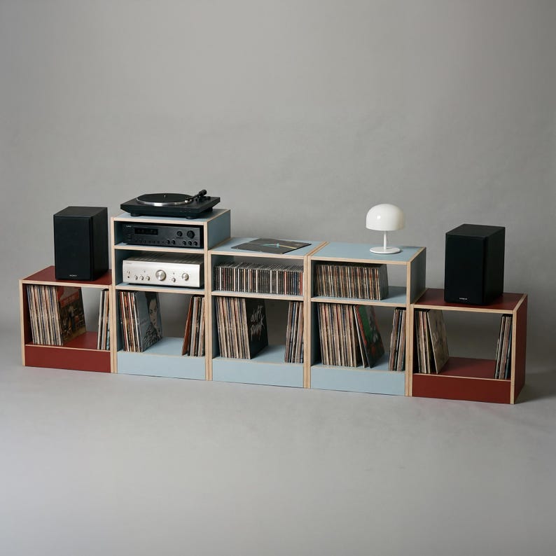 Modular plywood record player stand and vinyl storage system with natural plywood edges shown as a wide low console against a gray background. Oxide red and chalk blue modules hold a turntable and stereo components in the center with vinyl records and CDs stored across the shelves. Black speakers at both ends and a white table lamp show the scale and styling of the unit.