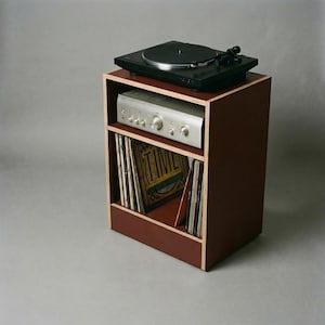 Modular plywood record player stand made from 18mm plywood in oxide red with natural plywood edges shown from a high front angled view against a gray background. A black turntable sits on top with a silver stereo component on the shelf below and vinyl records stored upright in the lower section.