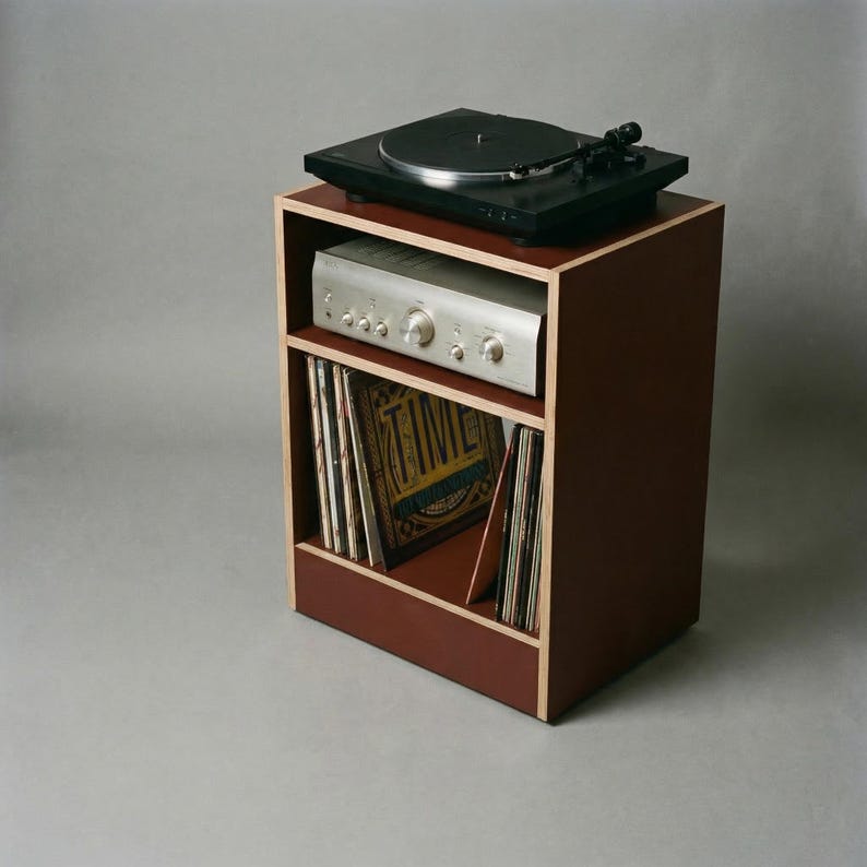 Modular plywood record player stand made from 18mm plywood in oxide red with natural plywood edges shown from a high front angled view against a gray background. A black turntable sits on top with a silver stereo component on the shelf below and vinyl records stored upright in the lower section.