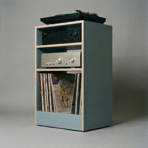 Modular plywood record player stand made from 18mm plywood in chalk blue with natural plywood edges shown from a front angled view against a gray background. A black turntable sits on top with two stereo components on the shelves below and vinyl records stored upright in the lower section.