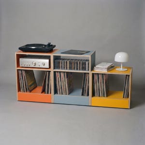 May include: A modular storage unit consisting of three stacked wooden cubes in orange, blue, and yellow. The unit holds a black turntable, a silver audio receiver, rows of vinyl records, a stack of books, and a small white mushroom-shaped lamp.