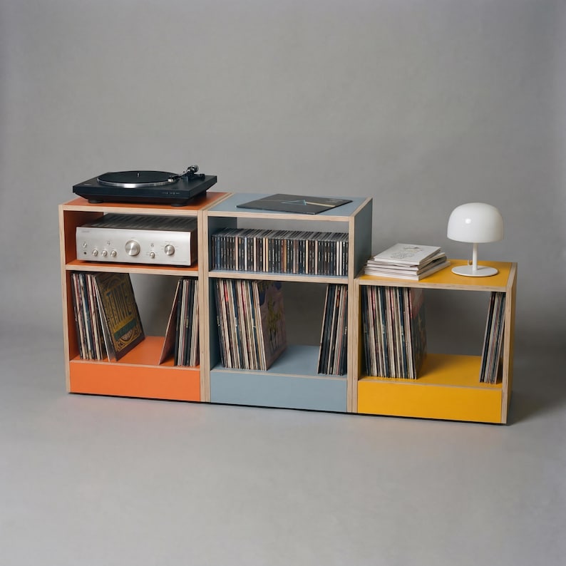 May include: A modular storage unit consisting of three stacked wooden cubes in orange, blue, and yellow. The unit holds a black turntable, a silver audio receiver, rows of vinyl records, a stack of books, and a small white mushroom-shaped lamp.