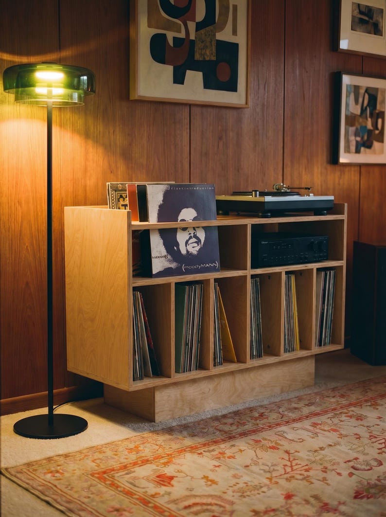 Groove No.3 birch plywood record player stand with vinyl storage, turntable, amplifier shelf, and records in a warm wood-paneled listening room.
