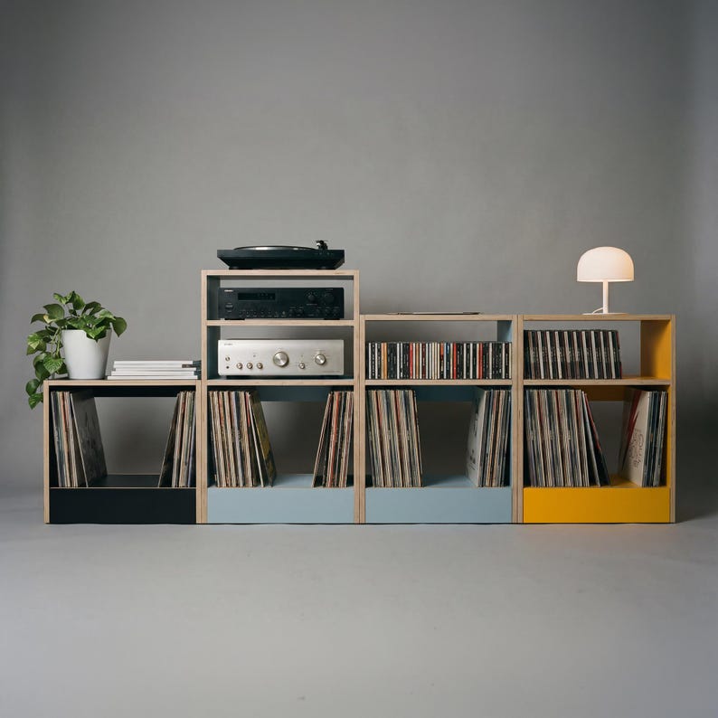Modular plywood record player stand configured as a wide low storage system with natural plywood edges and black chalk blue and yellow sections. A turntable and stereo components sit in the center while vinyl records and CDs are stored across the shelves. A white lamp and potted plant frame the setup to show the scale and styling of the unit.
