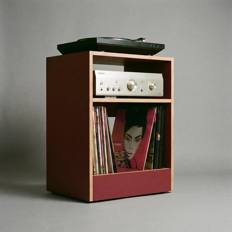 Modular plywood record player stand in oxide red with natural plywood edges shown from a front angled view against a gray background. A black turntable sits on top with a silver stereo component on the shelf below and vinyl records stored upright in the lower section.