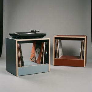 Modular plywood record player stand and vinyl storage units with natural plywood edges are shown as two separate pieces against a gray background. The blue unit on the left holds a black turntable on top and upright records below. The oxide red unit on the right adds extra vinyl storage and shows the flexible modular layout.
