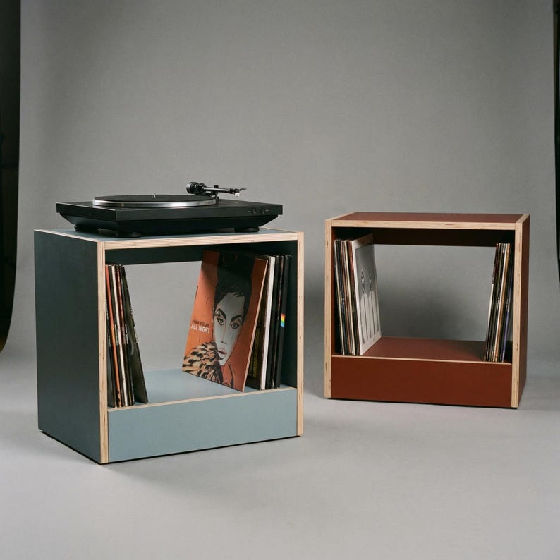 Modular plywood record player stand and vinyl storage units with natural plywood edges are shown as two separate pieces against a gray background. The blue unit on the left holds a black turntable on top and upright records below. The oxide red unit on the right adds extra vinyl storage and shows the flexible modular layout.
