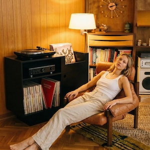 Puede incluir: Una mujer con una camiseta sin mangas blanca y pantalones beige se relaja en un sillón de cuero marrón. Un tocadiscos negro y equipo estéreo están sobre un mueble negro. Los discos de vinilo están apilados cerca, y una lámpara proporciona una iluminación suave.