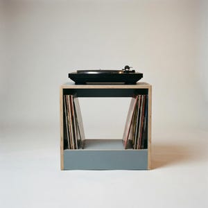 Modular plywood record player stand and vinyl storage unit with natural plywood edges shown from the front against a gray background. A black turntable sits on top. Vinyl records are stored upright below in front of a chalk blue panel and the open shelf layout highlights the compact modular design.