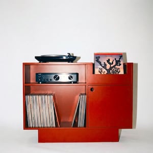 Puede incluir: Un soporte rojo de madera para tocadiscos con un tocadiscos negro, un amplificador y discos de vinilo. El soporte tiene un compartimento para guardar discos y un espacio para mostrar la carátula del álbum. La carátula del álbum en el soporte dice "DJ Koze".
