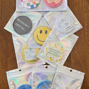 May include: A collection of round, colorful buttons with positive affirmations and designs, packaged in clear, iridescent pouches. The buttons feature phrases like "Be kind," "You're enough," and "Believe in yourself."