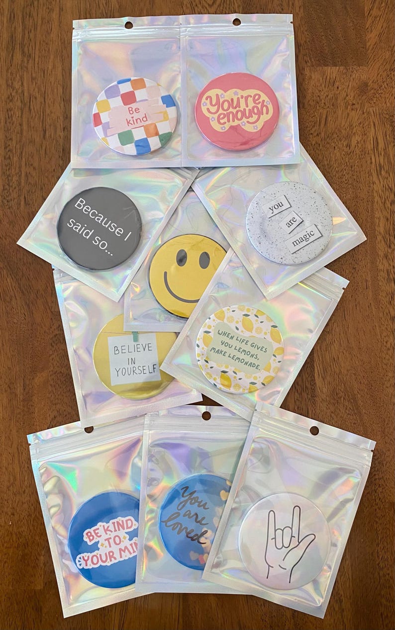 May include: A collection of round, colorful buttons with positive affirmations and designs, packaged in clear, iridescent pouches. The buttons feature phrases like "Be kind," "You're enough," and "Believe in yourself."