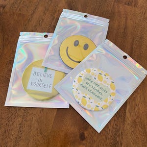 May include: Three round, yellow decorative items in clear, iridescent pouches. One has a smiley face, another reads "BELIEVE IN YOURSELF", and the third says "WHEN LIFE GIVES YOU LEMONS, MAKE LEMONADE."