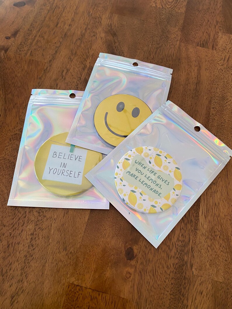 May include: Three round, yellow decorative items in clear, iridescent pouches. One has a smiley face, another reads "BELIEVE IN YOURSELF", and the third says "WHEN LIFE GIVES YOU LEMONS, MAKE LEMONADE."