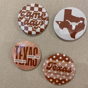 May include: Four circular pin-back buttons with Texas-themed designs. One features a brown and white checkerboard pattern with the text 'game day'. Another shows a brown Texas state silhouette with a white longhorn. Two others feature 'Texas' text.