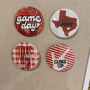May include: Four circular pinback buttons with red and white designs. One features a red glitter background with the text game day. Another shows a red map of Texas with the text Saturdays in LBK. A third has a red gingham pattern with Texas Tech Wreck 'Em Raiders text. The fourth has red stripes and a hand gesture graphic with the text Guns Up.