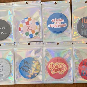 May include: A collection of round phone grips in clear plastic packaging. The grips feature various designs and text, including a hand gesture, the phrase "Because I said so...", a colorful pattern, and inspirational quotes. The grips are in a variety of colors.