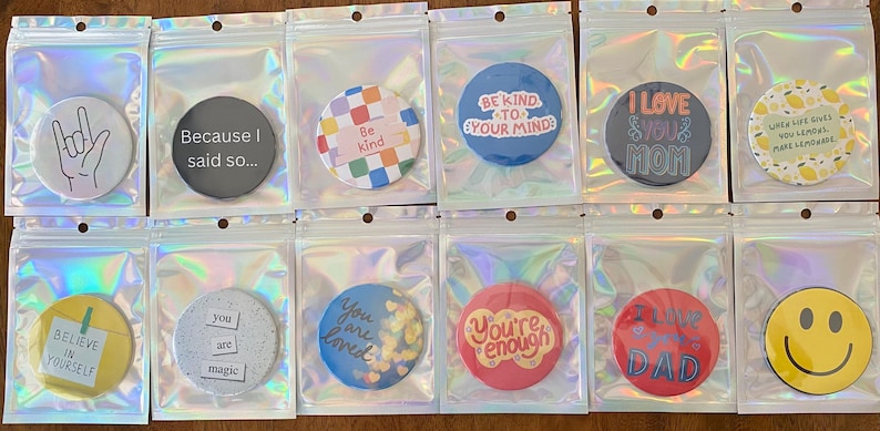 May include: A collection of round phone grips in clear plastic packaging. The grips feature various designs and text, including a hand gesture, the phrase "Because I said so...", a colorful pattern, and inspirational quotes. The grips are in a variety of colors.