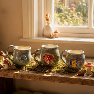 Op de afbeelding: Drie keramische mokken met grillige ontwerpen staan op een houten plank. De mokken zijn blauwgroen met unieke handvatten en hebben een paddenstoel, een beer en een wervelontwerp. Decoratieve paddenstoelen en eikels zijn rond de mokken verspreid.