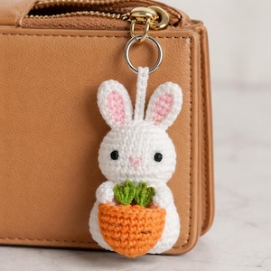 Puede incluir: Un llavero de conejo blanco tejido a ganchillo con orejas internas rosas y ojos negros, sosteniendo una zanahoria naranja con hojas verdes. El conejo está unido a un llavero plateado, colgando de una cartera de cuero marrón.