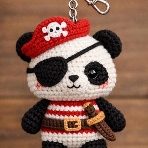 Pode incluir: Chaveiro de urso panda de crochê vestido de pirata. O panda usa um chapéu de pirata vermelho com uma caveira branca, tapa-olho preto e camisa listrada em vermelho e branco. Possui cinto marrom com fivela dourada e uma espada pequena.