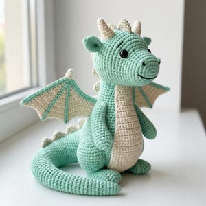 Puede incluir: Un juguete de dragón tejido a ganchillo en tonos verde menta y crema. El dragón tiene ojos grandes y amigables, cuernos pequeños y alas detalladas. El peluche hecho a mano está sobre una superficie blanca.