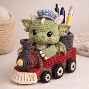 Peut inclure: Un dragon vert crocheté portant un chapeau rayé bleu et blanc, assis dans une locomotive rouge. Le train a des roues noires et un phare jaune. Le dragon tient un crayon et des stylos dans un récipient noir.