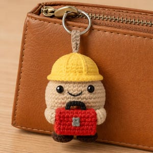 May include: A crocheted keychain featuring a smiling character wearing a yellow construction hat and holding a red briefcase. The keychain is attached to a silver ring and a tan strap. The background includes a brown leather wallet.