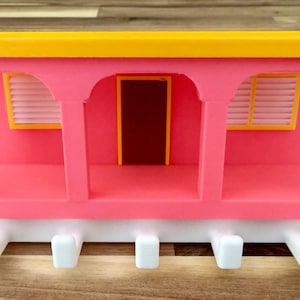 Op de afbeelding: Een roze en gele decoratieve wandorganizer in de vorm van een treinwagon. Het heeft boogvormige openingen, een bruine deur en witte haken. De organizer is ongeveer 30 cm lang.