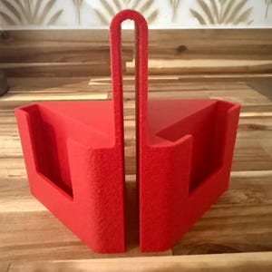 May include: A bright red, triangular-shaped object with a handle. The object appears to be a tool or container, possibly for holding or organizing items. The handle is a curved, U-shape, and the object has a textured surface.