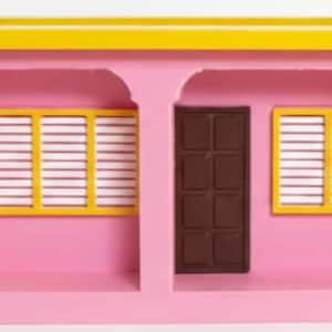 Op de afbeelding: Een roze miniatuurhuismodel met een geel dak en raamkozijnen. Het heeft een bruine deur en twee ramen met witte luiken. Het ontwerp is eenvoudig en kleurrijk, geschikt voor decoratieve doeleinden.